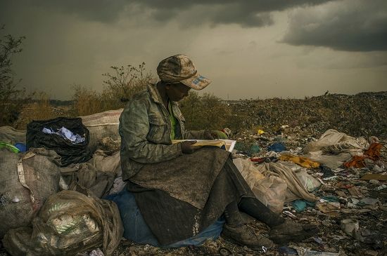 当代热点类单幅一等奖《在Dandora垃圾场》：在肯尼亚奈洛比，一个捡垃圾的妇女坐在30英亩大的垃圾场中，静静看着她从垃圾场中捡到的书。照片有种油画般的诗意。这个妇女最大的愿望就是在捡拾垃圾之余有更多的时间去看看书，哪怕只是工业书籍的目录。(美国摄影师Micah Albert拍摄)
